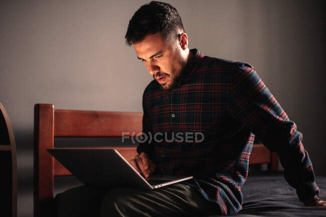 Концентрований чоловік використовує ноутбук, сидячи на ліжку вдома — стокове фото