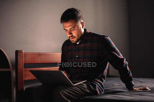 Hombre usando ordenador portátil sentado en la cama en casa - foto de stock