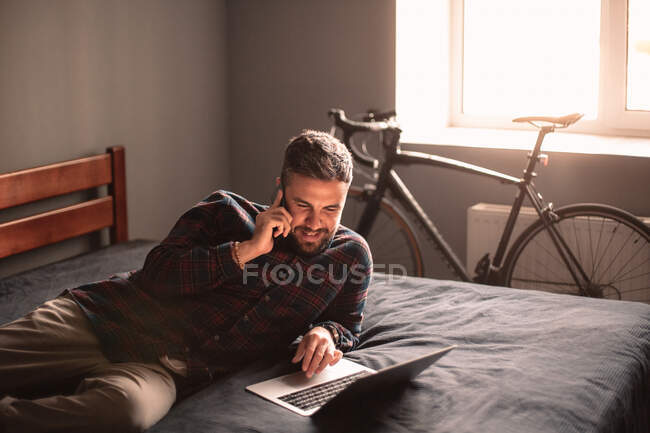 Glücklicher Geschäftsmann, der zu Hause mit dem Laptop auf dem Smartphone spricht — Stockfoto