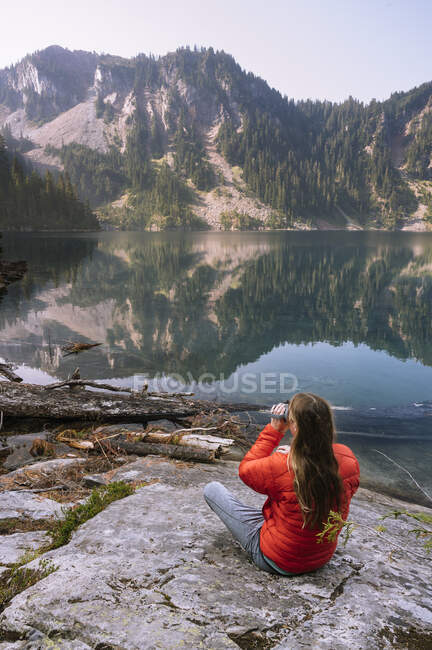 Joven sentado en el lago de la montaña, mirando a la distancia - foto de stock