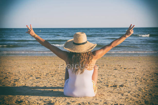 Beautiful woman in hat sitting on sandy beach, back view — Stock Photo