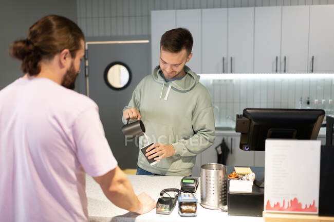 Man in hoodie pouring milk from pitcher into cup while preparing coffee for male client — Stock Photo