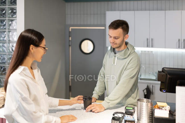 Uomo in felpa con cappuccio che dà bevanda calda alla donna asiatica mentre lavora nella moderna caffetteria — Foto stock