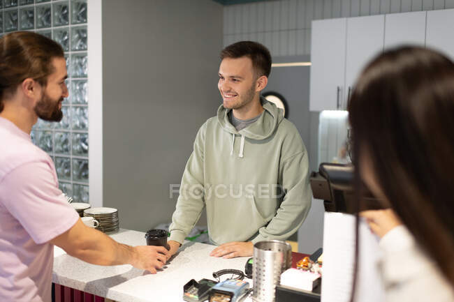 Homem em roupas casuais sorrindo e dando café para ir ao cliente masculino no café moderno — Fotografia de Stock