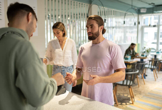 Бородатий чоловічий клієнт розмовляє з баристами, готуючи каву під час очікування замовлення — стокове фото