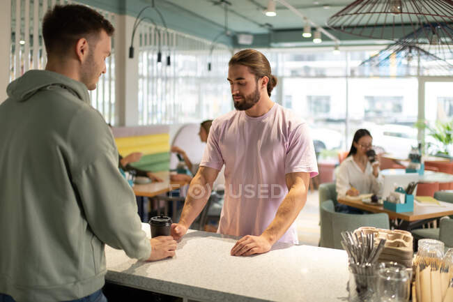 Bearded male client taking coffee to go from barista during visit in modern cafe — Stock Photo