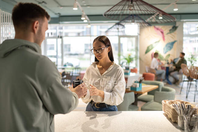 Mann in legerer Kleidung serviert asiatischer Frau während der Arbeit im Café Kaffee zum Mitnehmen — Stockfoto