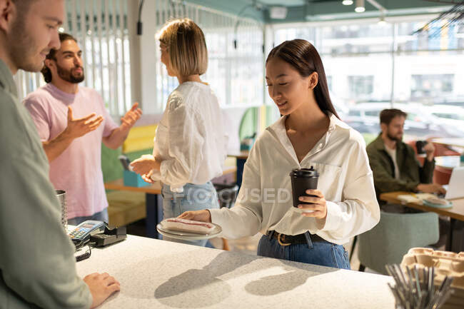 Asiática hembra con café para ir examinando postre mientras toma orden de barista - foto de stock