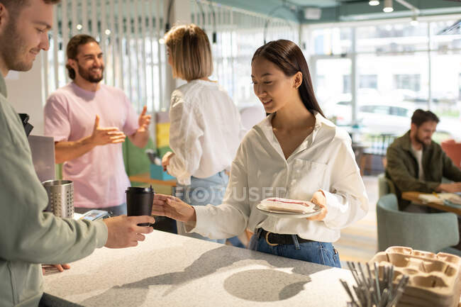 Мужчина, подающий кофе на вынос и сладкий десерт радостной азиатке в современном кафе — стоковое фото