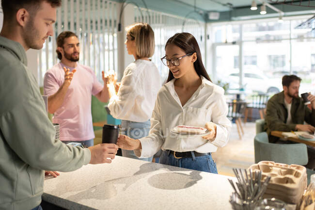 Glad Asian woman with dessert taking takeaway coffee from barista in modern cafe — Stock Photo