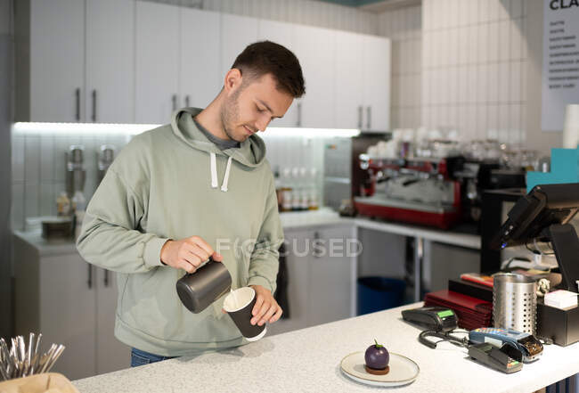 Мужчина-бариста в повседневной одежде наливает молоко в чашку для еды на вынос во время работы в кафе — стоковое фото