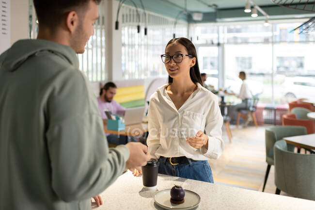 Varón barista y asiático cliente femenino hablando mientras se reúnen en la cafetería moderna - foto de stock