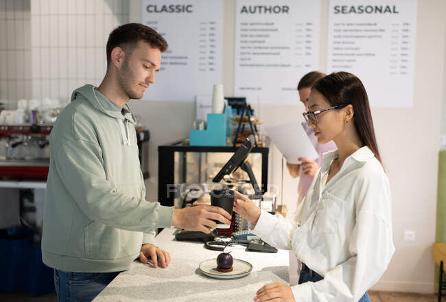 Mann in lässiger Kleidung serviert asiatischen Kunden im Kaffeehaus Heißgetränk und Dessert — Stockfoto