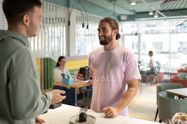 Male barista talking with male client with drink to go while working in coffee house — Stock Photo
