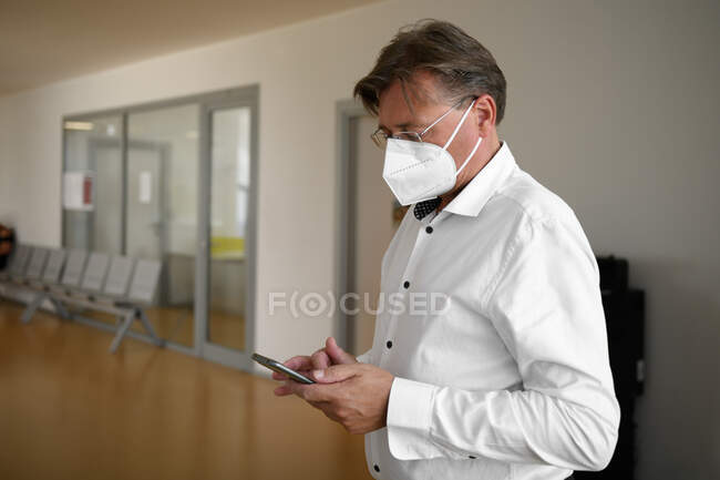 Homem adulto em óculos, usando máscara protetora contra doenças infecciosas transmissíveis e como proteção contra a gripe no escritório público, usando e olhando para o smartphone — Fotografia de Stock
