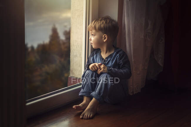 Niño pequeño con ropa azul sentado en el suelo por la ventana y el baño - foto de stock
