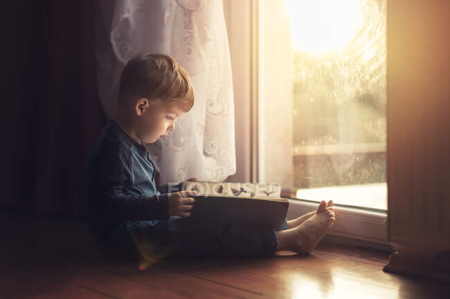 Vue latérale du petit garçon assis sur le sol par la fenêtre et la lecture — Photo de stock