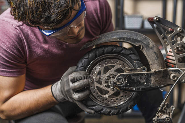 Ein sitzender Mechaniker mit Schutzbrille setzt das Rad eines Elektrorollers in der Werkstatt wieder ein. — Stockfoto