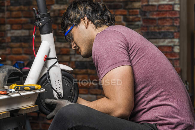 Un meccanico latino seduto mette una ruota di uno scooter elettrico al suo posto nella sua officina. — Foto stock