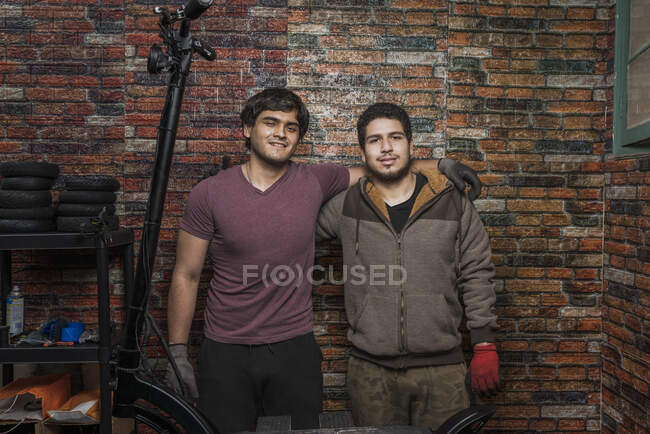 Frontansicht zweier Mechaniker-Kollegen, die sich umarmen und in die Kamera schauen, die in der Werkstatt vor einer Ziegelwand steht. — Stockfoto