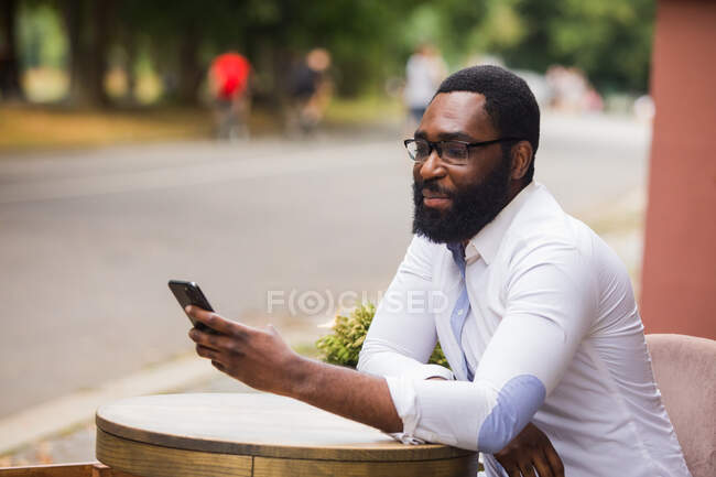 El joven con estilo está sentado en un café de la calle y utiliza un teléfono inteligente. Se endereza el pelo mientras hace una videollamada. - foto de stock