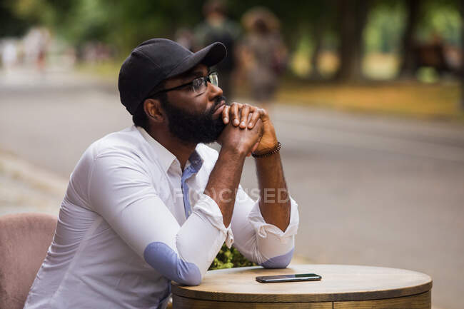 Retrato de um jovem que está sentado a uma mesa em um café de rua. Ele está vestindo uma camisa branca e boné — Fotografia de Stock