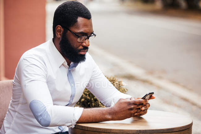 El joven está sentado en una mesa en un café de la calle y mirando en el teléfono inteligente. Lleva una camisa blanca de moda y una gorra. - foto de stock