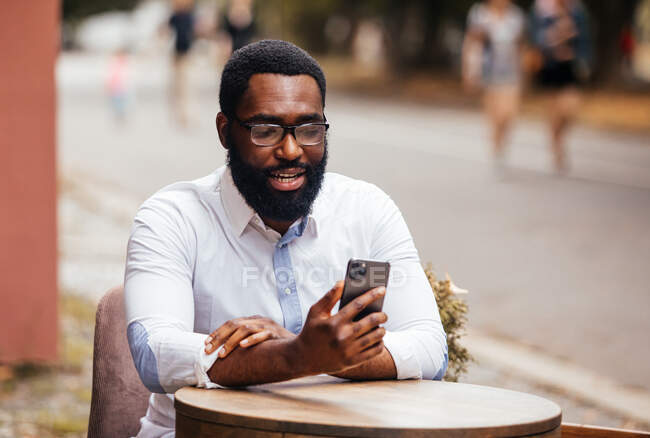 Der stylische junge Mann sitzt in einem Straßencafé und bedient sich eines Smartphones. Er glättet seine Haare bei einem Videoanruf — Stockfoto