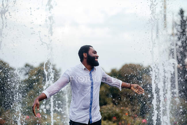 El joven sonriente se está divirtiendo en una fuente de la ciudad. Lleva una camisa blanca mojada. - foto de stock
