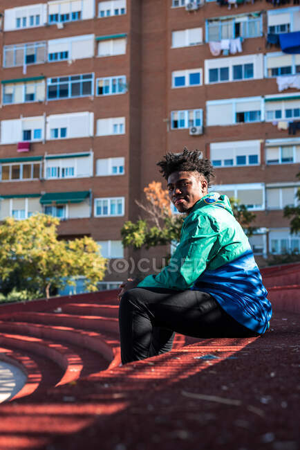 Ritratto di un giovane ragazzo nero seduto in città. Sfondo blocco di appartamenti. — Foto stock