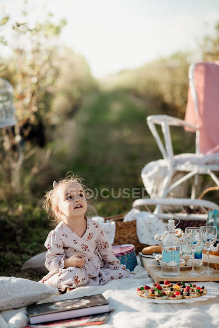 Menina sentada ao lado de um bolo em um pano de piquenique e olhando — Fotografia de Stock