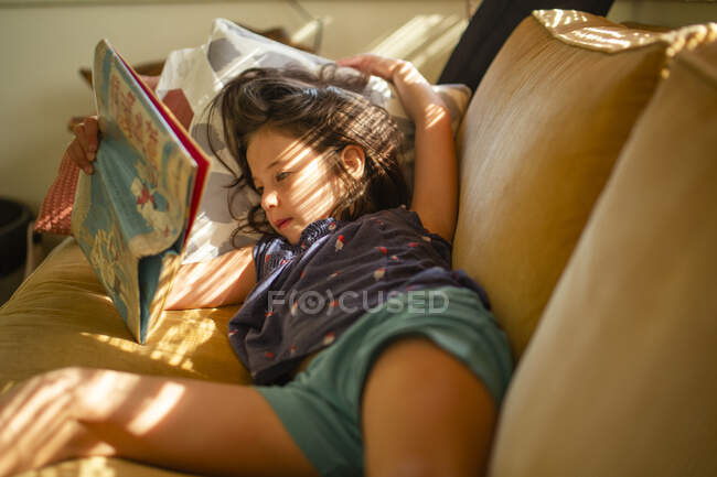 Little girl lying on sofa and reading book — Stock Photo