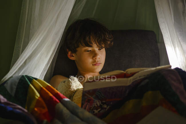 Kleiner Junge liegt mit Buch im Bett und blickt in Kamera — Stockfoto