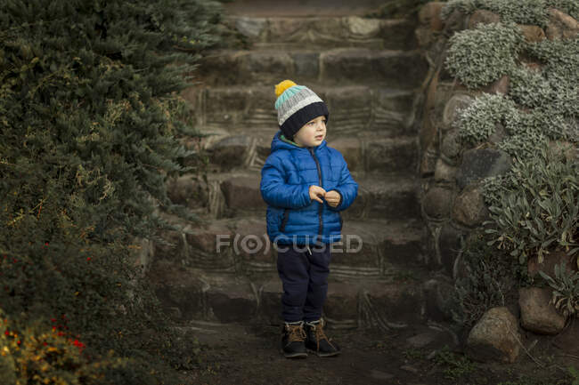 Menino de pé em casaco azul na frente de escadas de pedra no jardim — Fotografia de Stock