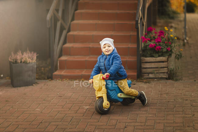 Menino de casaco azul montando moto de brinquedo ao lado da escadaria — Fotografia de Stock