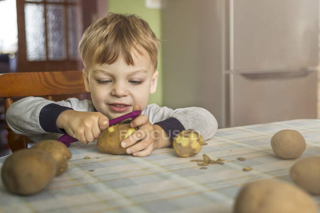 Small blonde boy learning how to peel potatoes with peeler sitti — Stock Photo