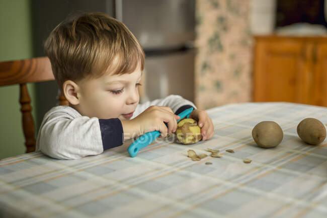 Pequeno menino loiro aprendendo a descascar batatas com descascador sitti — Fotografia de Stock