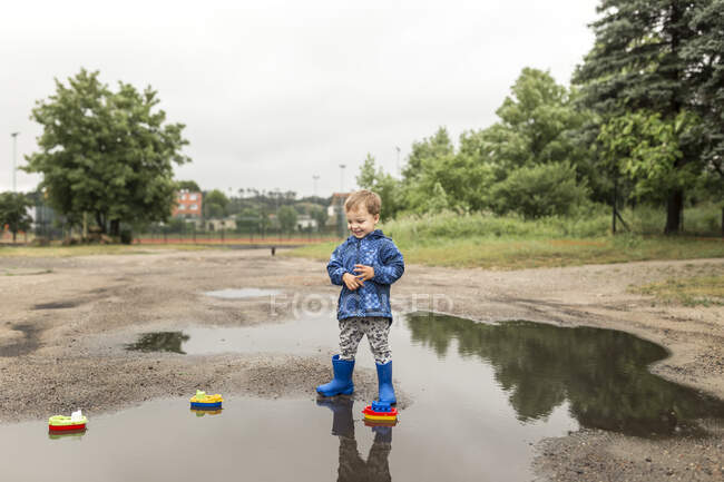 Menino loiro pequeno em casaco azul e azul wellies de pé e lo — Fotografia de Stock