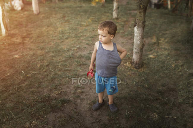 Маленький блондин стоит в саду в синих шортах и держит — стоковое фото