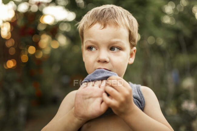 Portrait of a boy biting his summer top while standing in garden — Stock Photo