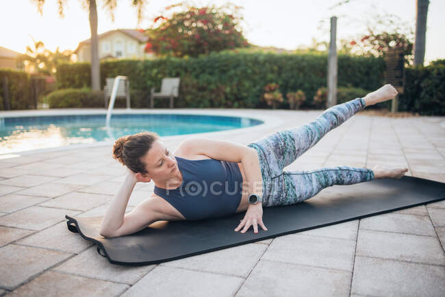 A woman doing mat pilates and leg raises next to a pool at sunrise — Stock Photo