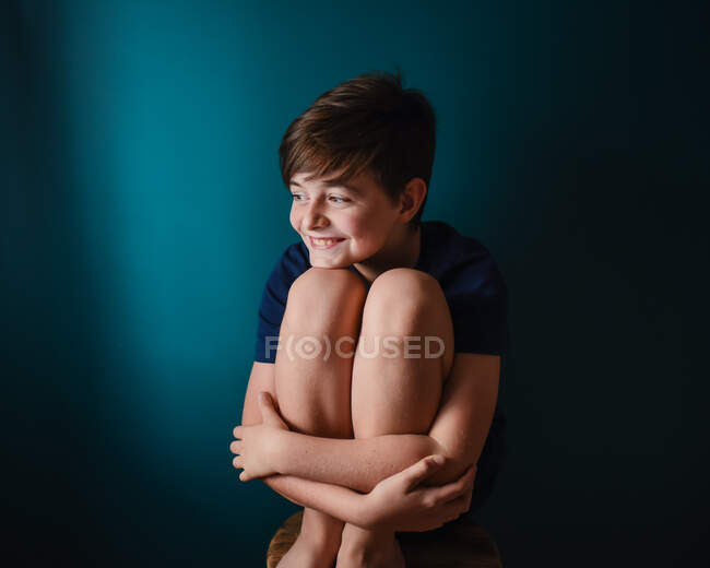 Niño feliz sentado en un taburete contra una pared azul oscuro. - foto de stock