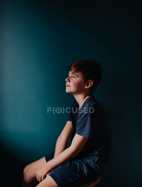 Jeune garçon réfléchi assis sur un tabouret contre un mur bleu foncé. — Photo de stock