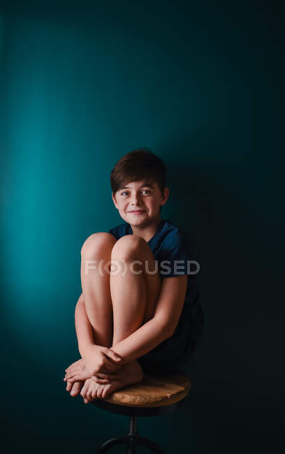 Glücklicher kleiner Junge sitzt auf einem Schemel vor einer dunkelblauen Wand. — Stockfoto