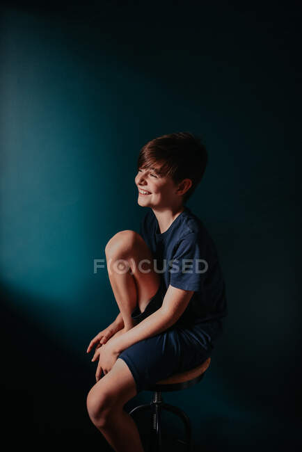 Щасливий хлопчик сидить на стільці біля темної блакитної стіни.. — стокове фото