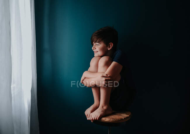 Happy young boy sitting on a stool against a dark blue wall. — Stock Photo