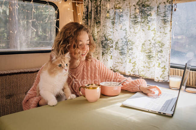 Woman and ginger cat in trailer are eating breakfast at laptop. — Stock Photo