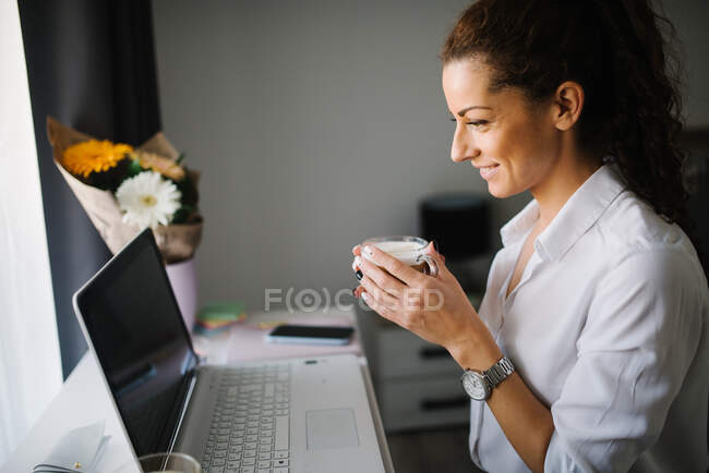 Mulher olhando para ele laptop e sorrindo enquanto segurando uma xícara de co — Fotografia de Stock