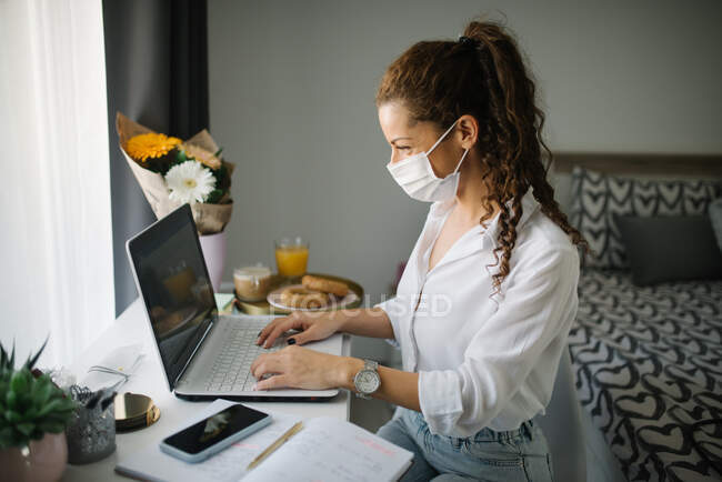 Mulher trabalhando em seu laptop com uma máscara protetora. — Fotografia de Stock