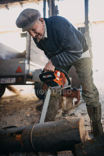 Старик улыбается, распиливая дрова бензопилой. — стоковое фото
